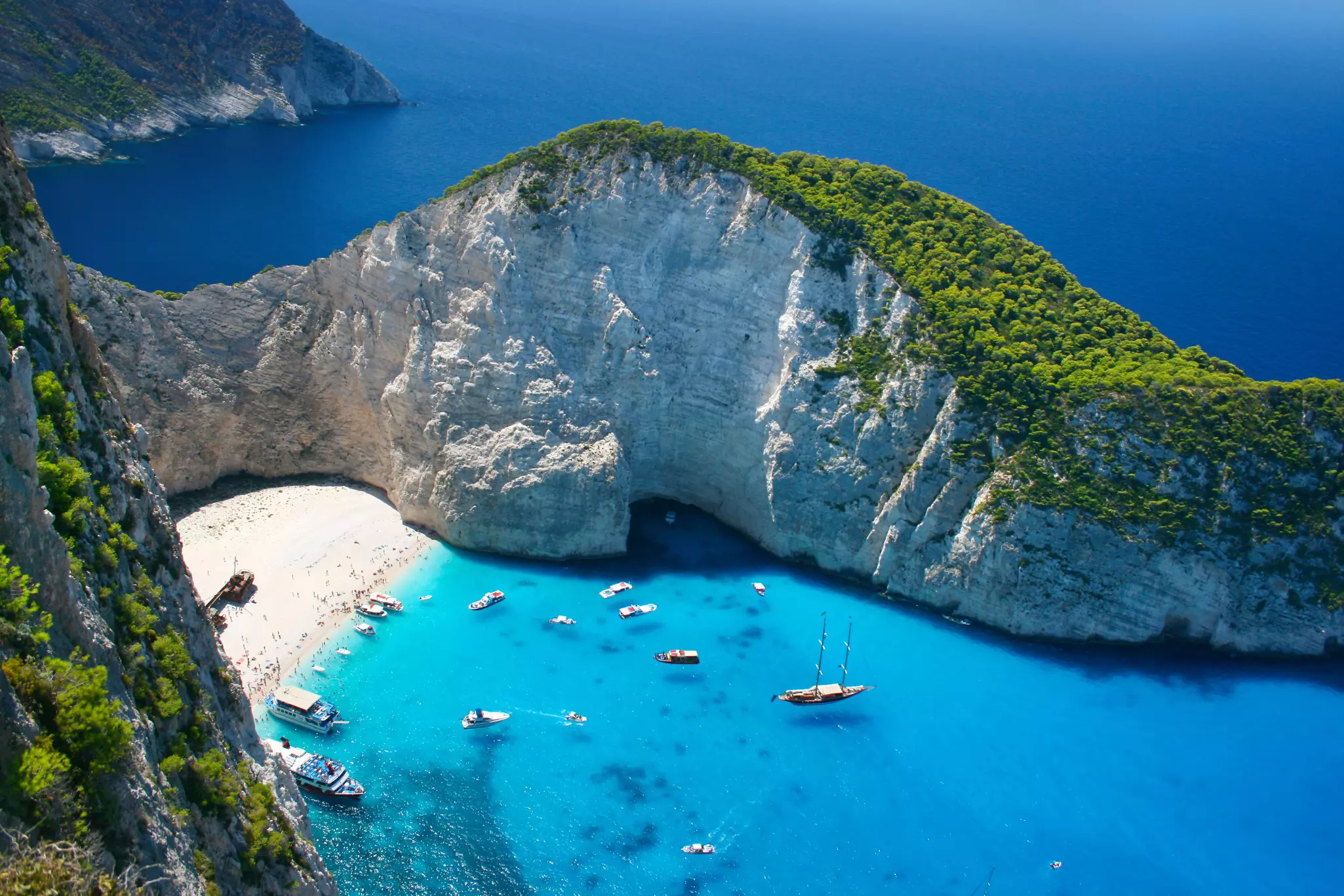 An aerial view of Navagio Beach, a sheltered cove in Greece with white sand, blue waters and a large wrecked ship on the shore.