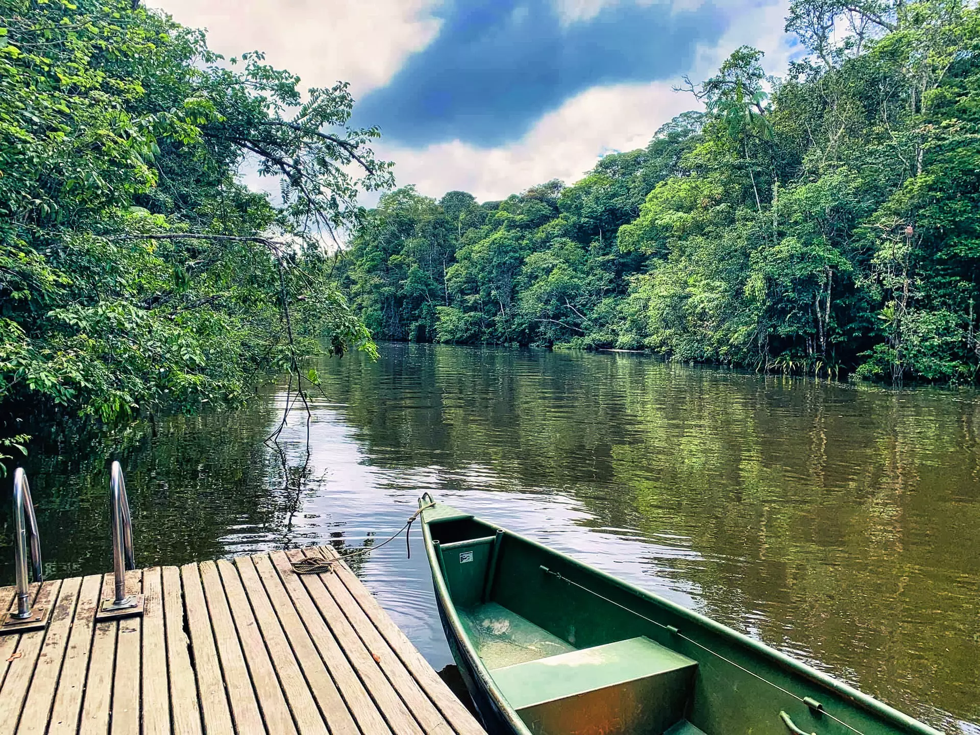 A small canoe near a wooden dock at the edge of a wide river surrounded by trees.