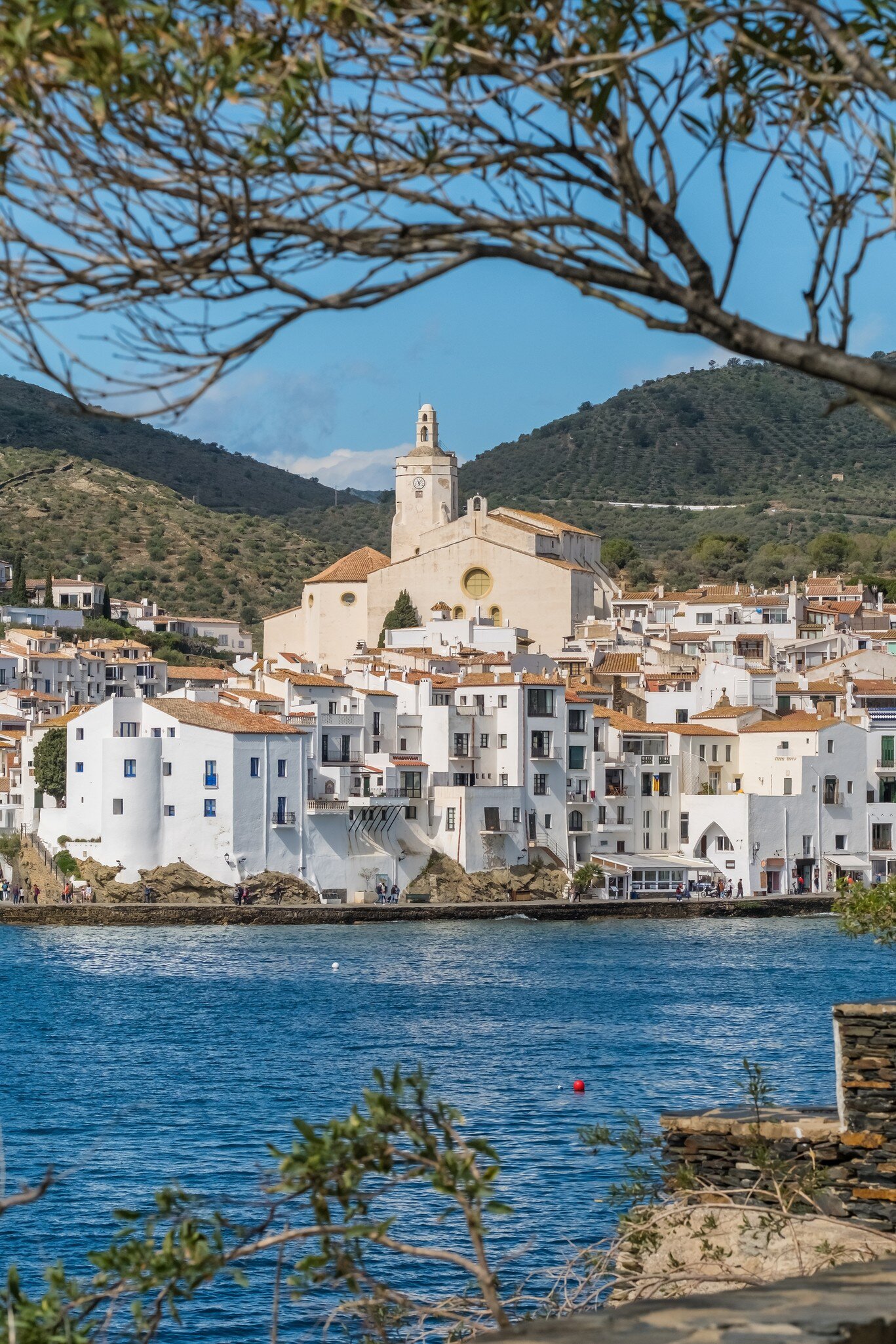 View of the town of Cadaqués, the Església de Santa María and the beautiful Costa Brava shore in Spain, framed by tree branches.