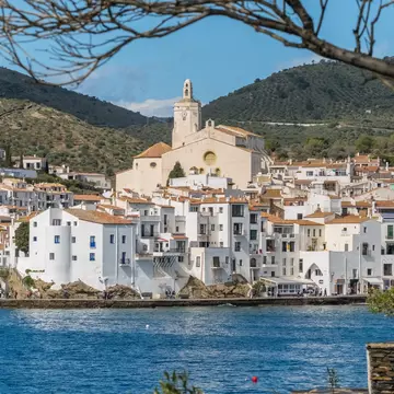 View of the town of Cadaqués, the Església de Santa María and the beautiful Costa Brava shore in Spain, framed by tree branches.