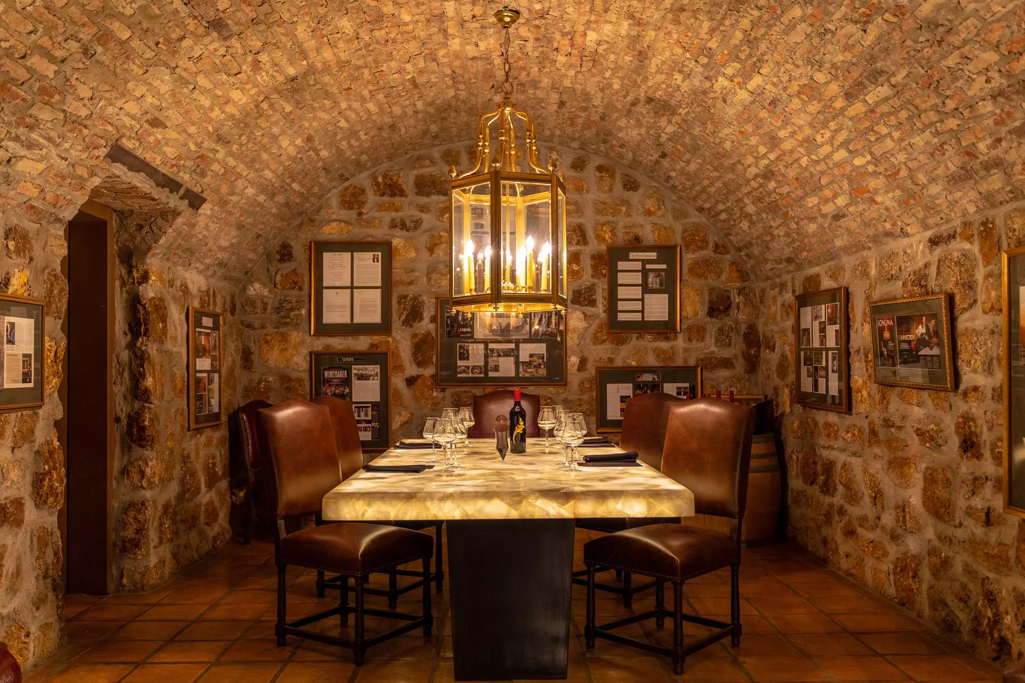 A room with stone walls has a table with place settings and wine glasses on top of it. Brown-cushioned chairs surround the table.