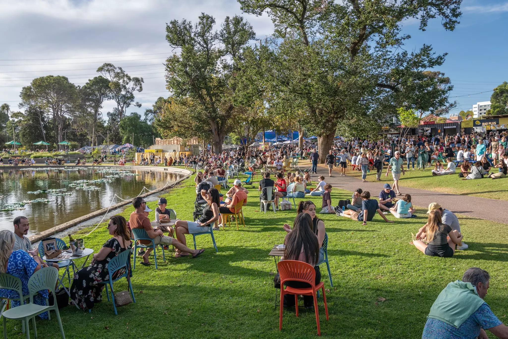People sit on the grass in the evening sun beside a large pond at a festival.
