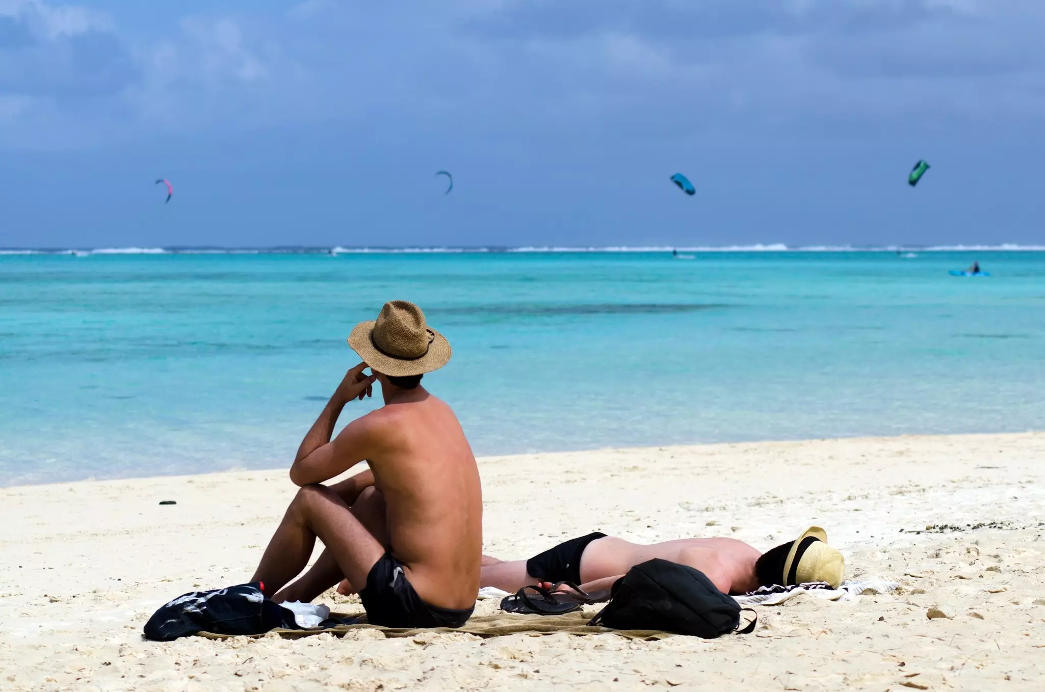 Two men in straw hats relax on a beach. Above the turquoise waters of the sea, kitsurfers are visible in the sky.