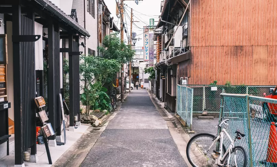 Traditional shops and houses lining a narrow street with a bike resting against a chainlink fence in the foreground