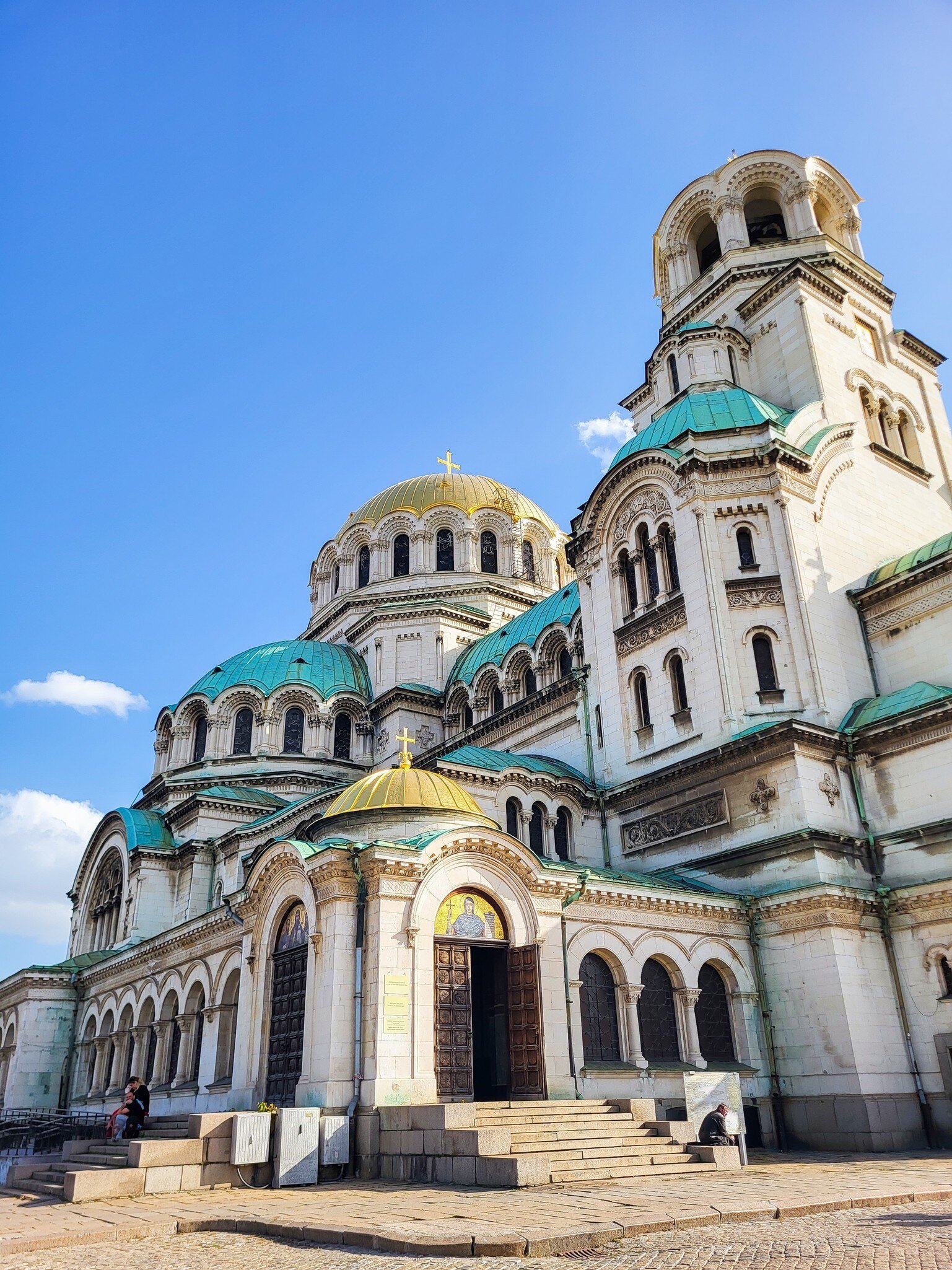 A white stone cathedral crowned by a gilded central dome and a cluster of green copper domes, with a mosaic above its bronze doors.