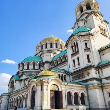 A white stone cathedral crowned by a gilded central dome and a cluster of green copper domes, with a mosaic above its bronze doors.