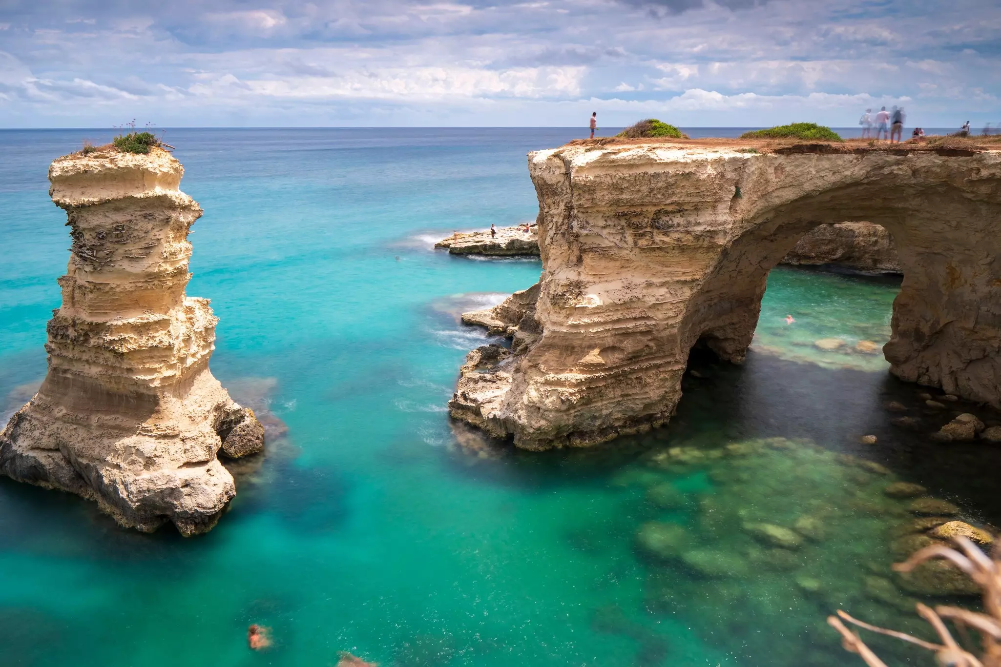People stand on a vast natural rock arch out at sea, while others swim close to nearby rock stacks.