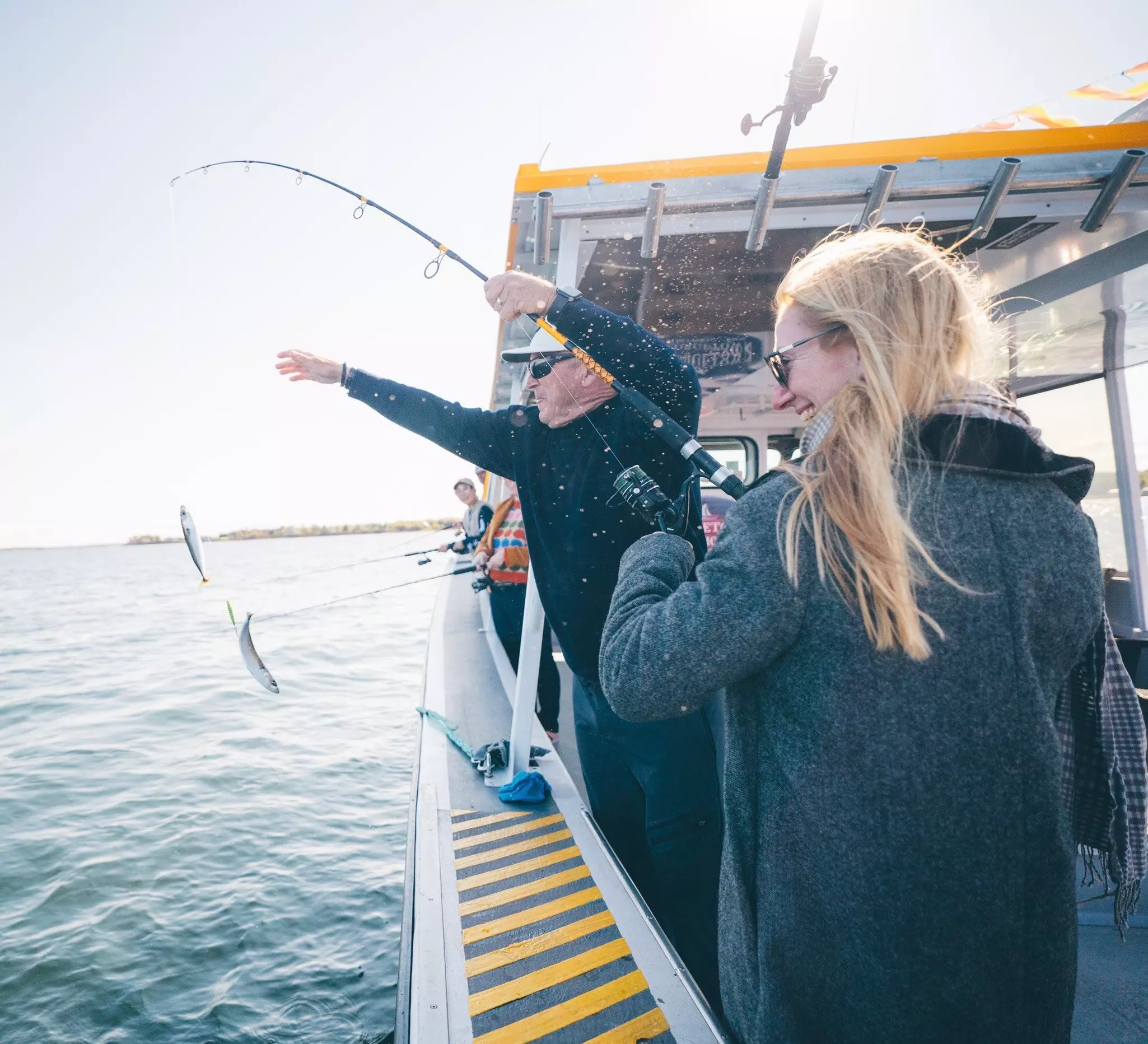 Two people standing on the deck of a boat pull up small fish caught on a line.