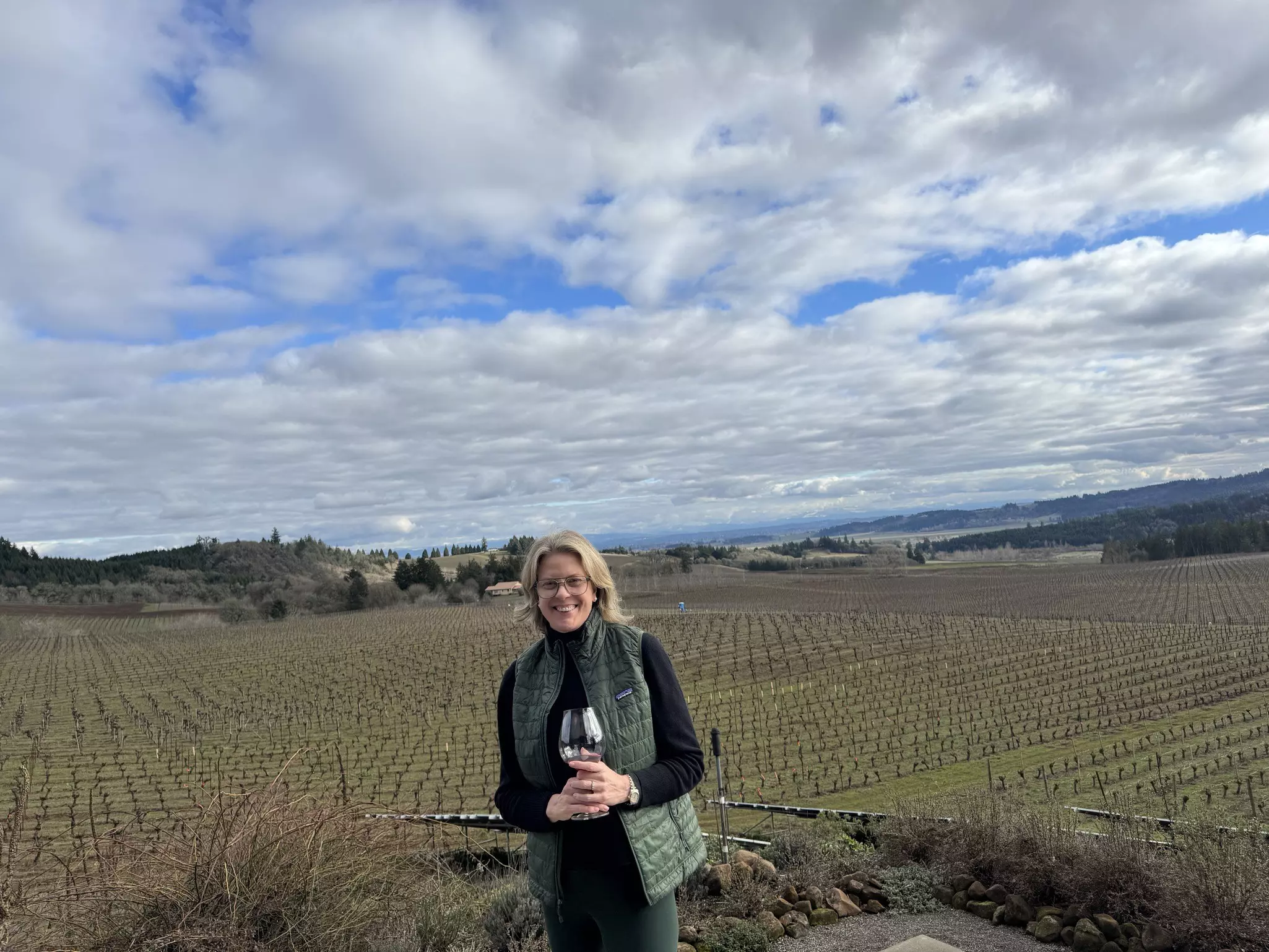 A woman with light hair and glasses, wearing a black shirt and green puffer vest, smiles and holds a wine glass while standing in front of a dormant vineyard with hills in the distances and clouds in the sky on an otherwise sunny day.