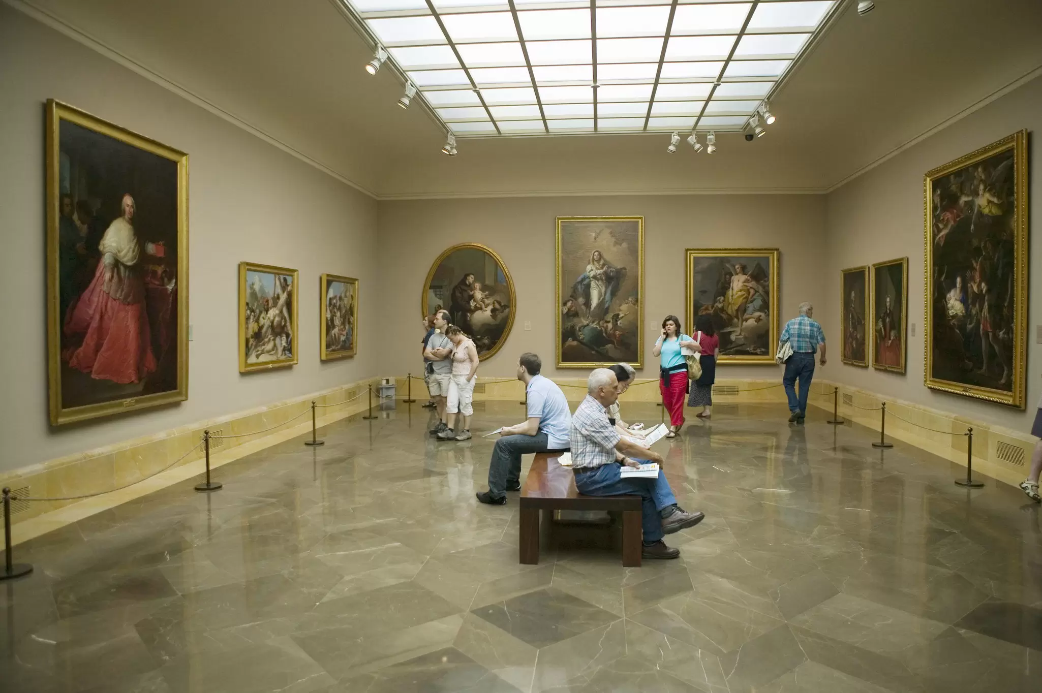 People sit on a central bench while others walk around a gallery lined with large paintings