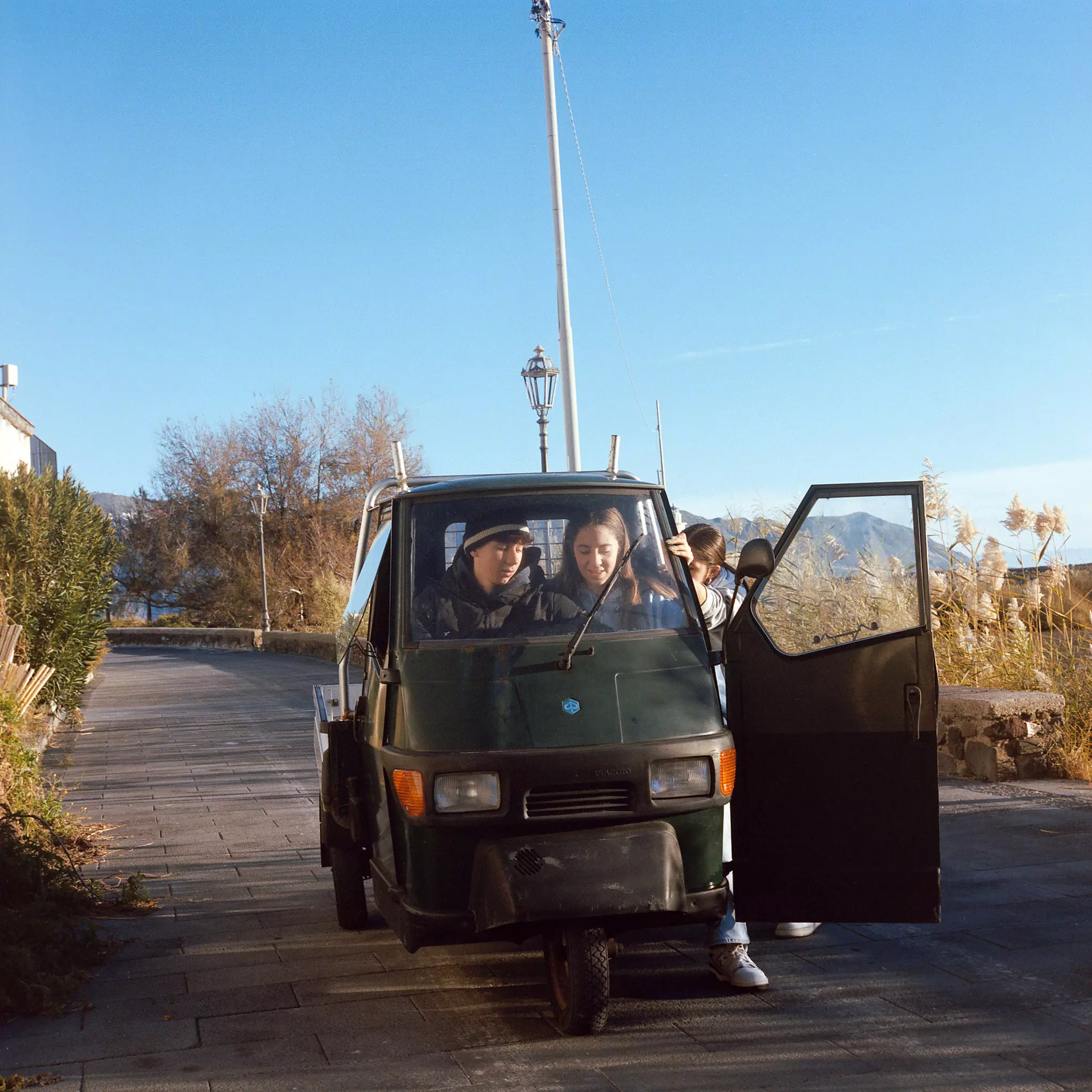 Two young people sit in a small-three wheeled vehicle parked on a small road in a village.