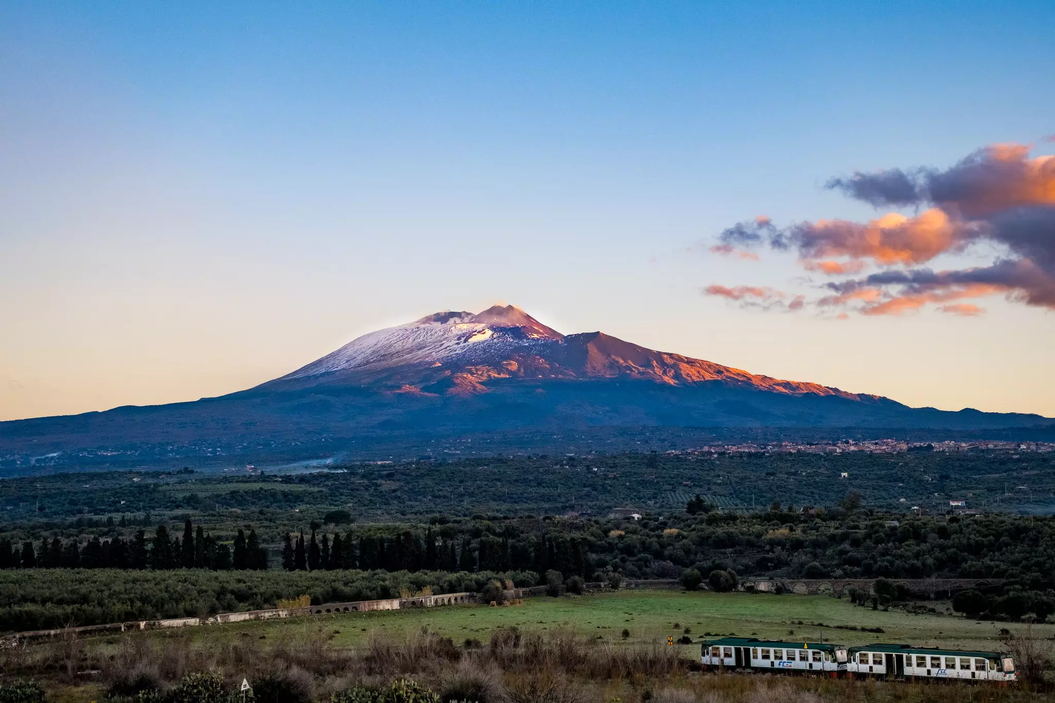 A train in the foreground crosses a green field in the morning; a massive crater is in the background.