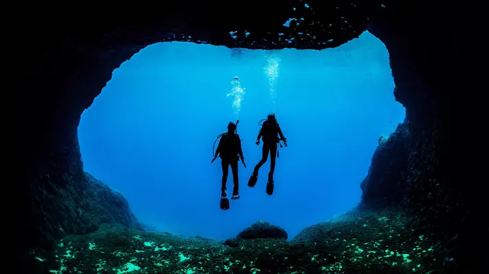 Silhouettes of two people in scuba gear underwater at the opening of a cave.