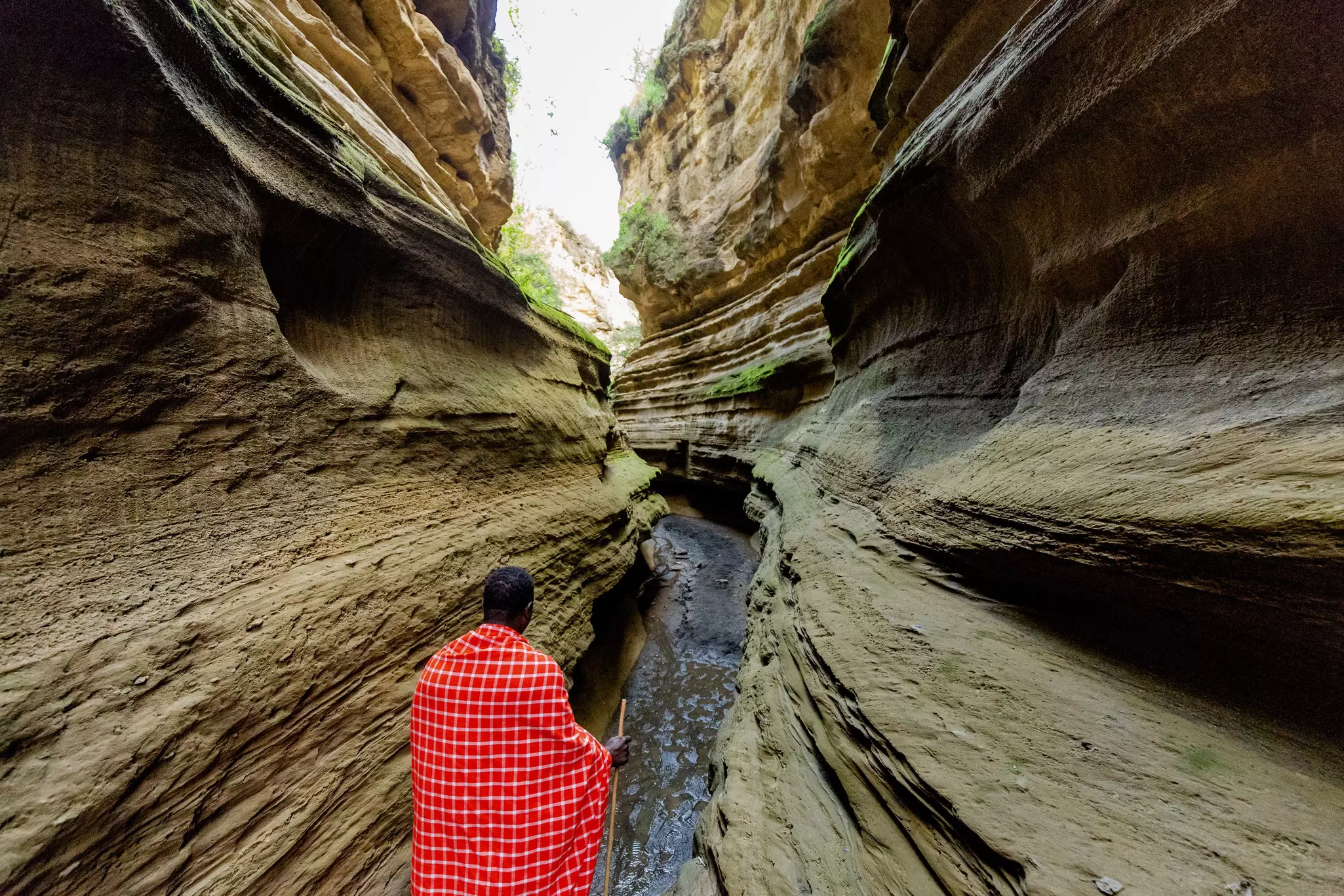 A man in a red cape-like covering carries a stick as he walks through a narrow path between two large rock formations that is partially filled with water