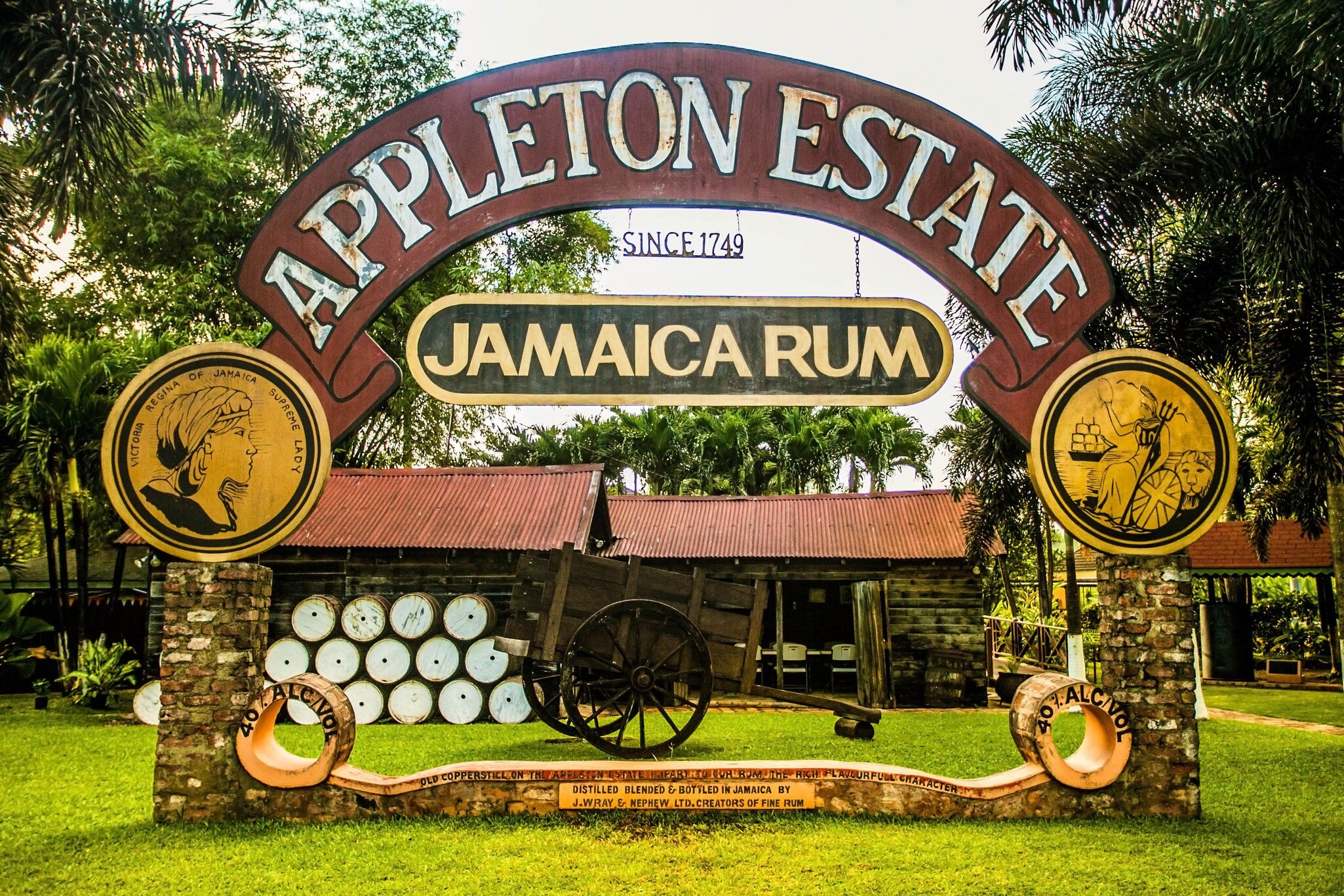 A painted archway sign above stacked barrels, a wooden cart and a tin-roofed shed on a lawn.
