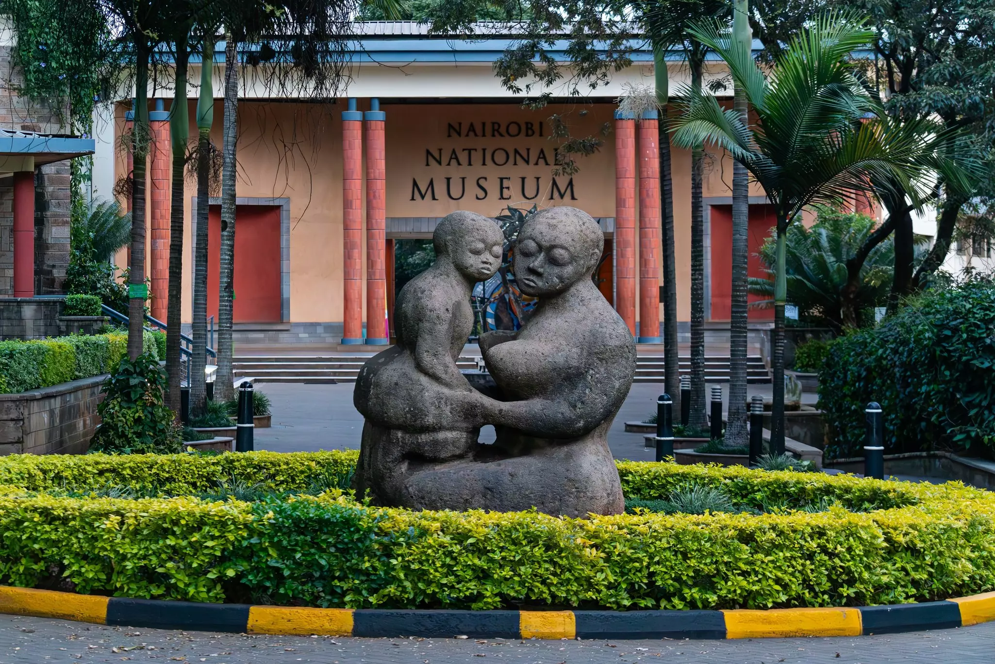 A sculpture depicting a mother and child is seen on a planted area in front of the entrance to a museum.