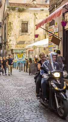 People ride a scooter on a narrow street in Italy.