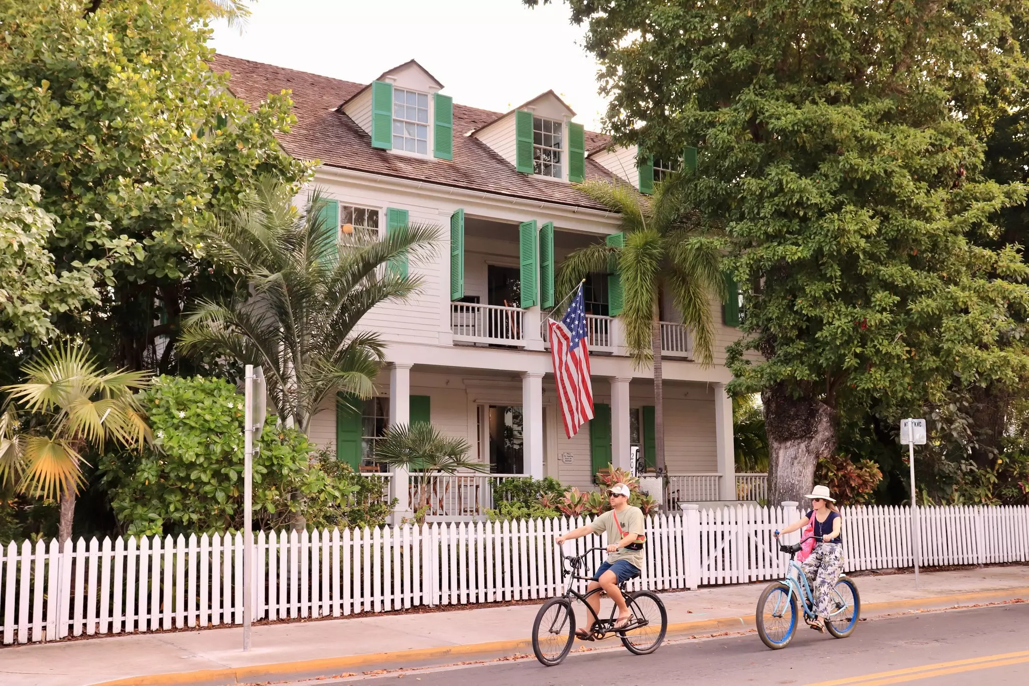 A man and a woman ride bikes in front of a white picket fence with a white house with green shutters behind