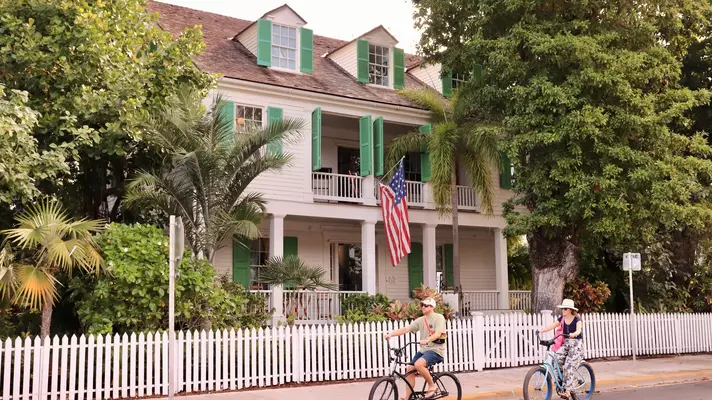 A man and a woman ride bikes in front of a white picket fence with a white house with green shutters behind