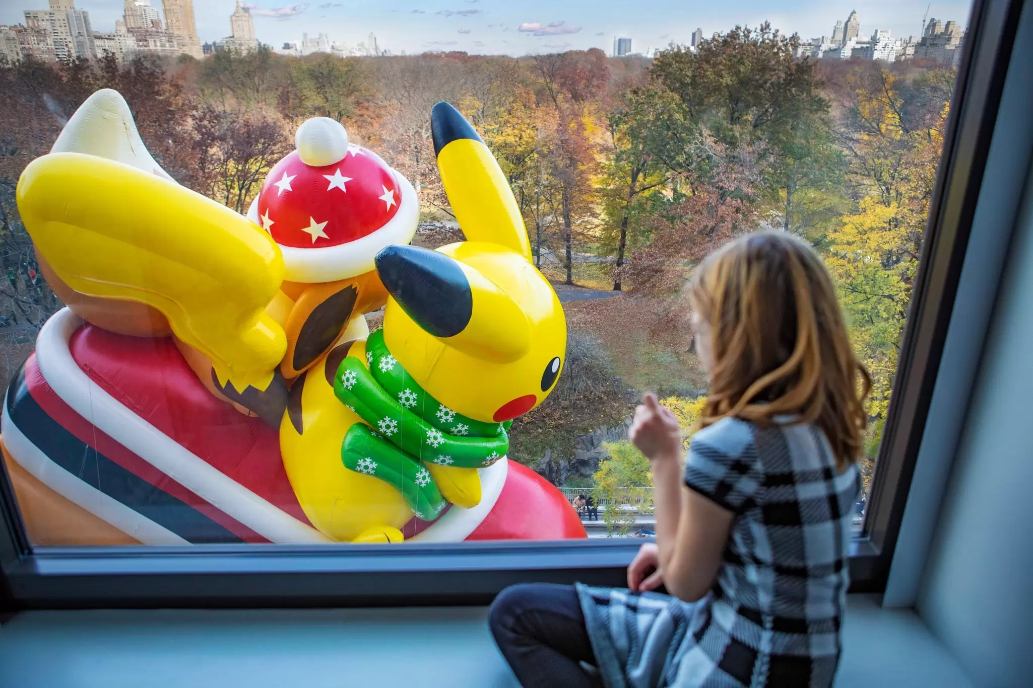 A child watches a large yellow balloon character (Pikachu) pass by a hotel room window: TWatching the Macy’s Thanksgiving Day Parade. Marriott International