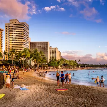 People on a sandy beach and in the water at sunset; tall buildings line the shore.