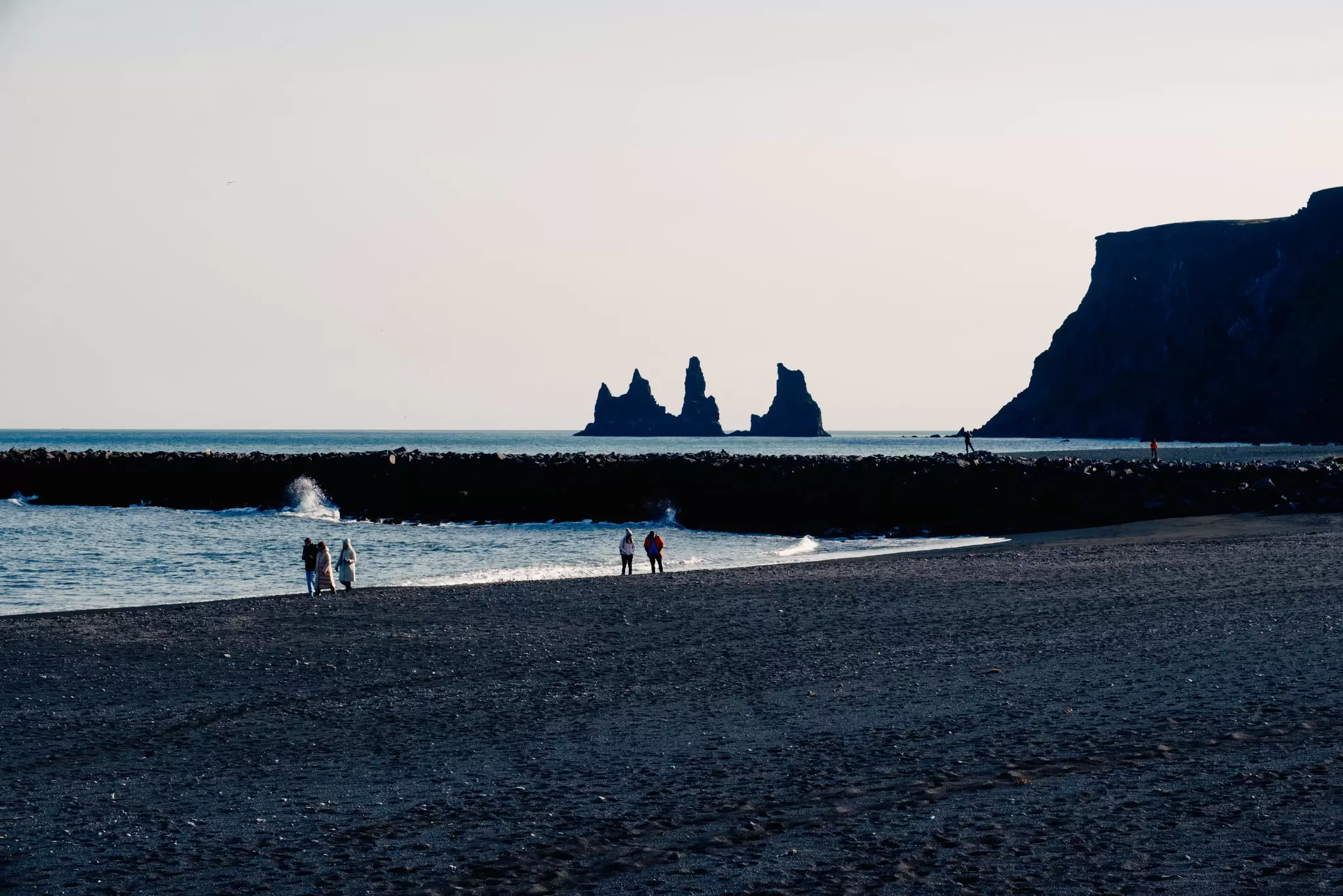 People stand on a black-sand beach with giant rock formations coming out of the water in the distance