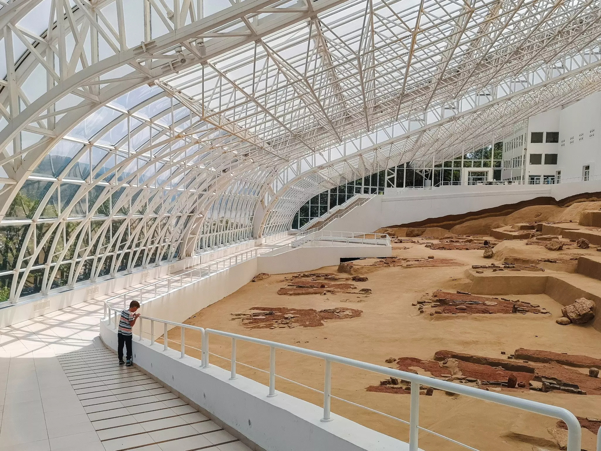 A child peers over the side of a railing to look at the remnants of an ancient fishing village at an archaeological site protected by a glass and steel building.