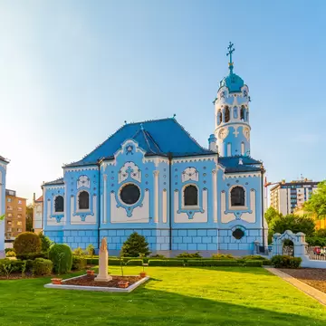 Powder-blue church with ornate white trim, a blue tiled roof and a round clock tower beside a lawn.