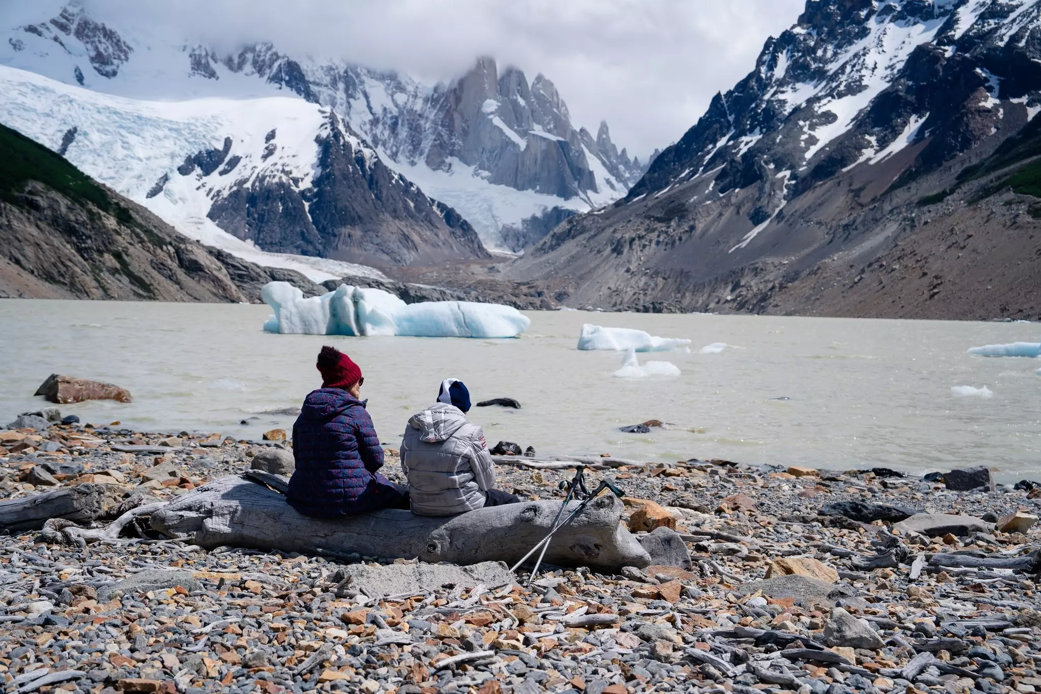 Two hikers sit on a weathered log on the rocky shore by a lake with glacier fragments floating in it; there are snow-covered mountains in the background.