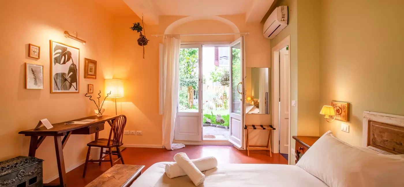 A hotel bedroom with warm lighting, a bed, a desk and a chair, and French doors opening out onto a garden