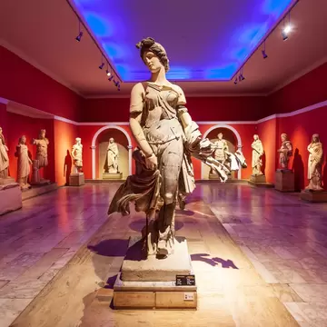 A deep red gallery hall lined with white marble statues on plinths under spotlights, their shadows thrown across the wall.