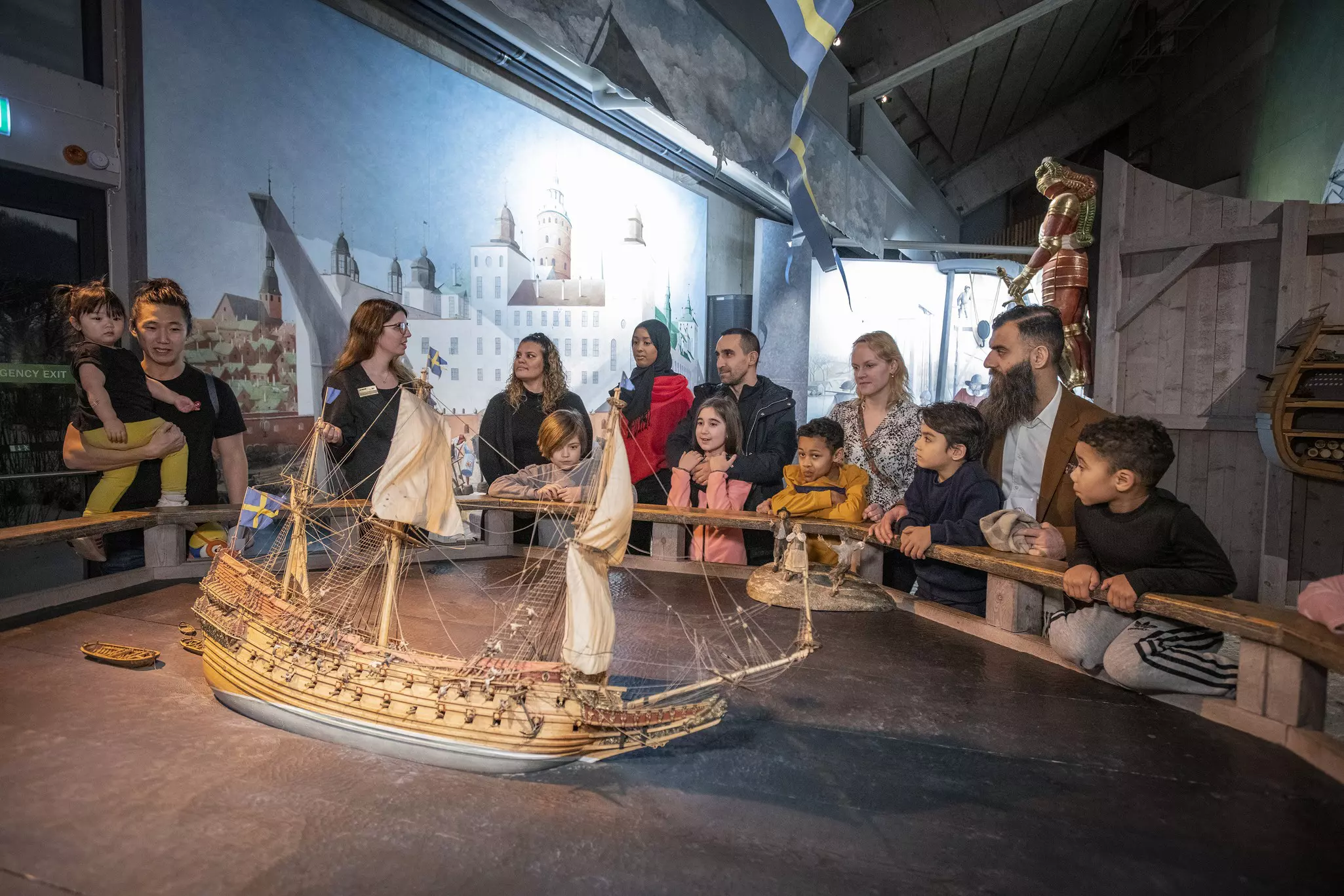 A multi-ethnic group of tourists gather round a model of a ship and listen to a museum guide at Stockholm's Vasa Museum