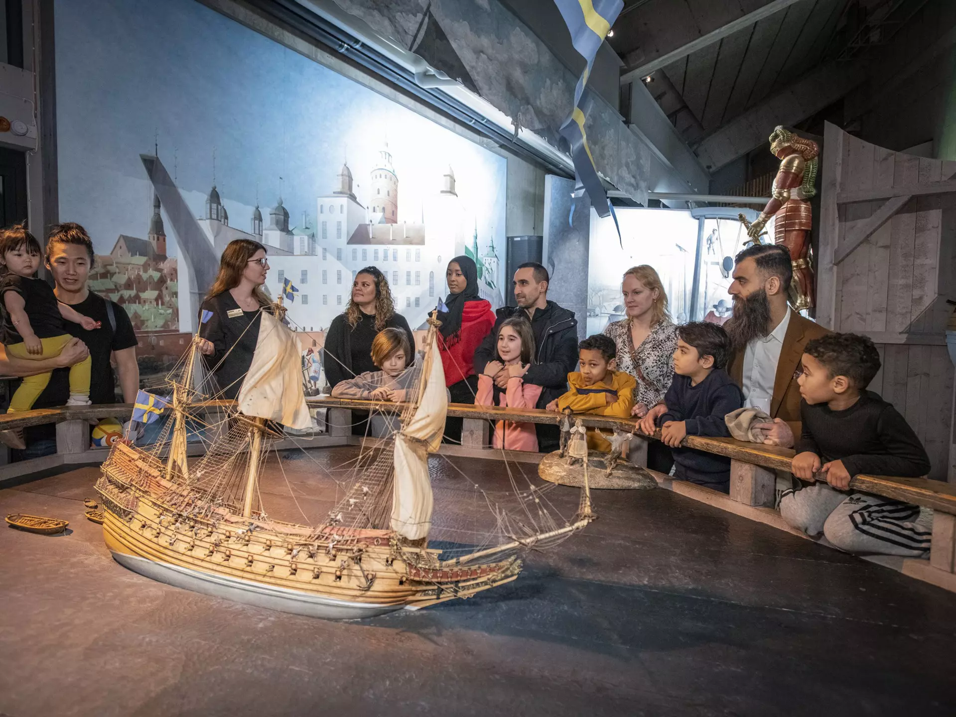 A multi-ethnic group of tourists gather round a model of a ship and listen to a museum guide at Stockholm's Vasa Museum