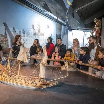 A multi-ethnic group of tourists gather round a model of a ship and listen to a museum guide at Stockholm's Vasa Museum