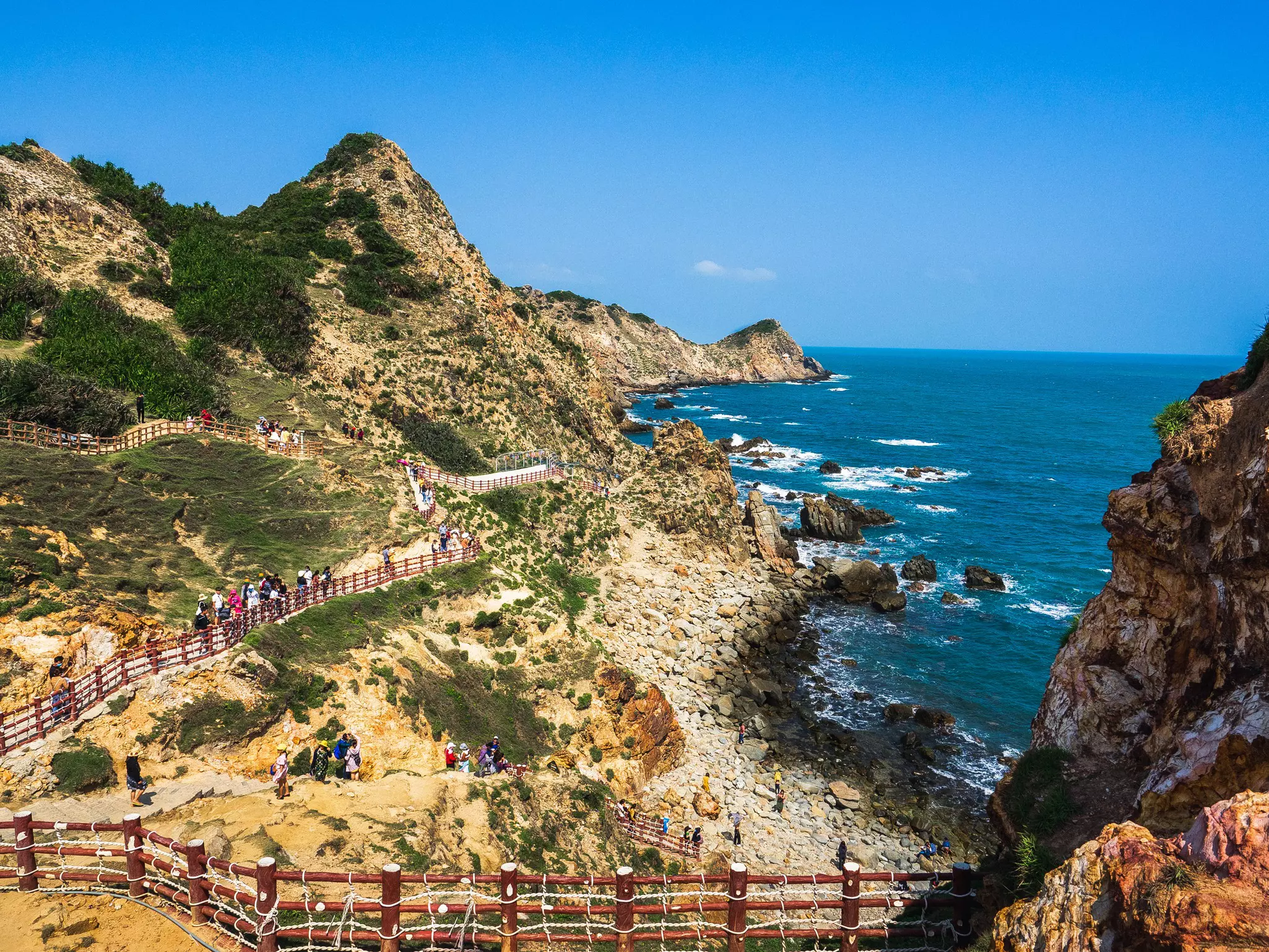 People walk along a pathway set in the side of cliffs to reach a rocky shoreline.