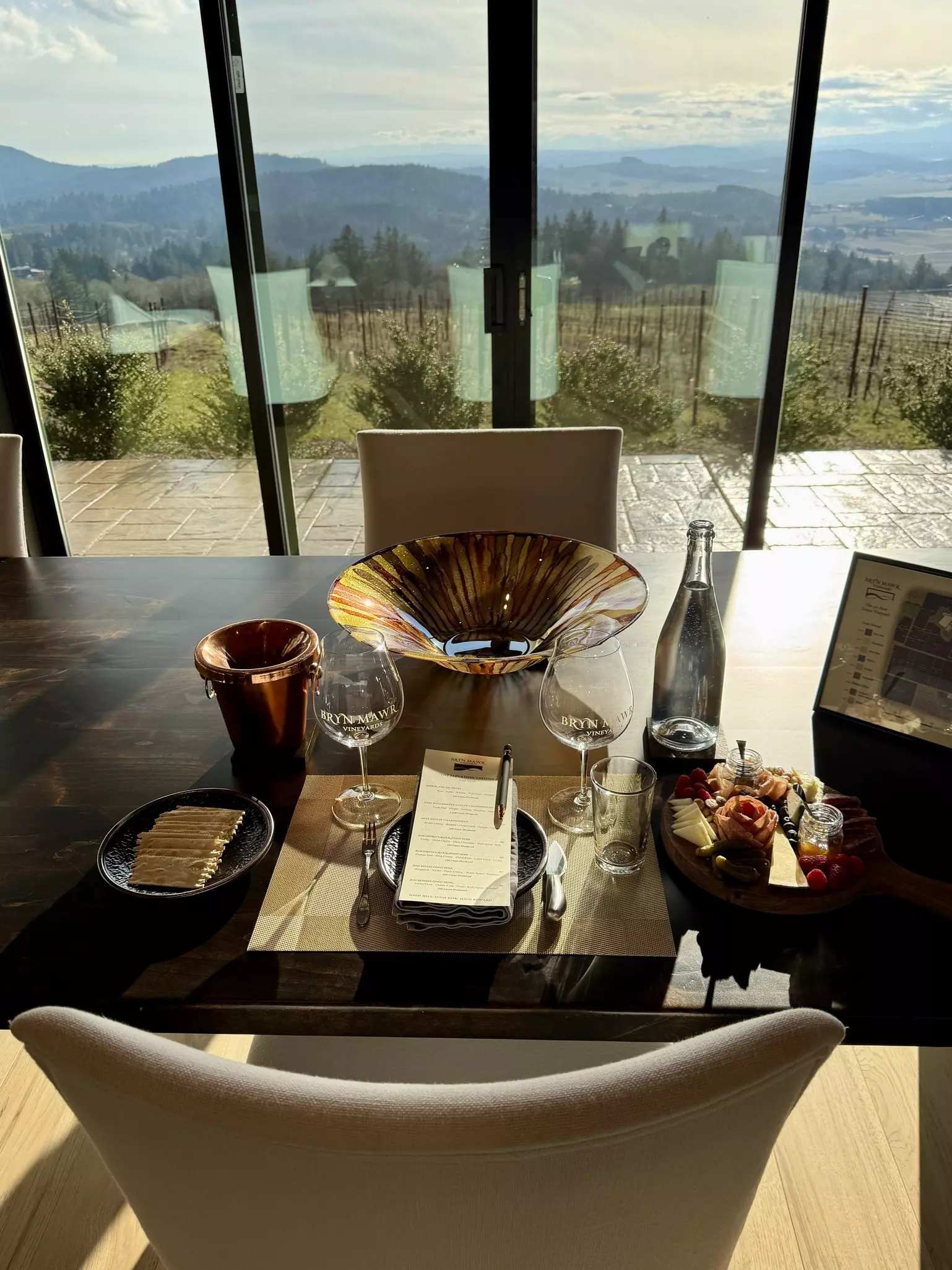 A plate of crackers and another with cheese and jams border a restaurant table setup with menu and wine glasses. Beyond, low forested hills can be seen through floor-to-ceiling windows on a sunny day.