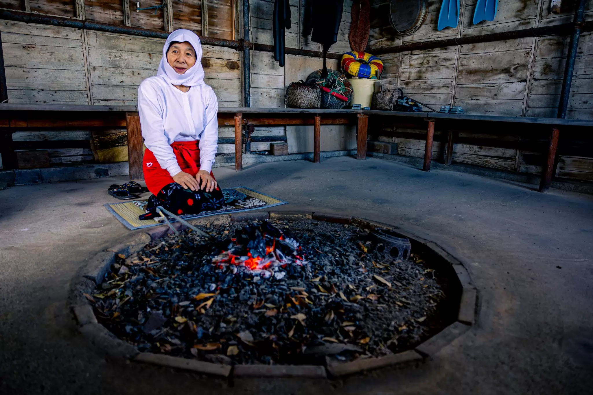 A diver in uniform sits beside the coals of a fire inside a small wooden hut.
