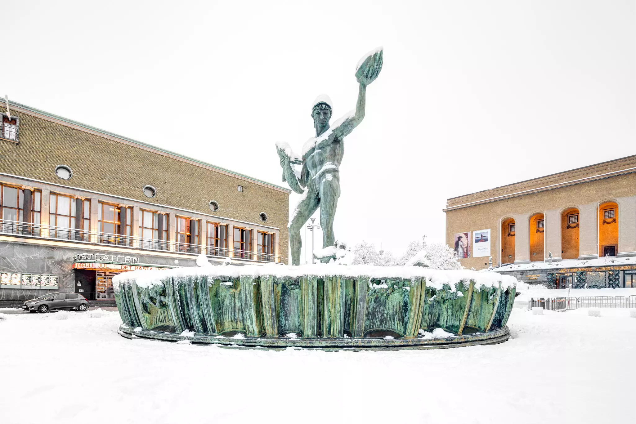 A bronze statue of a naked Greek god graces a fountain in a city square. Snow covers the fountain and the plaza.