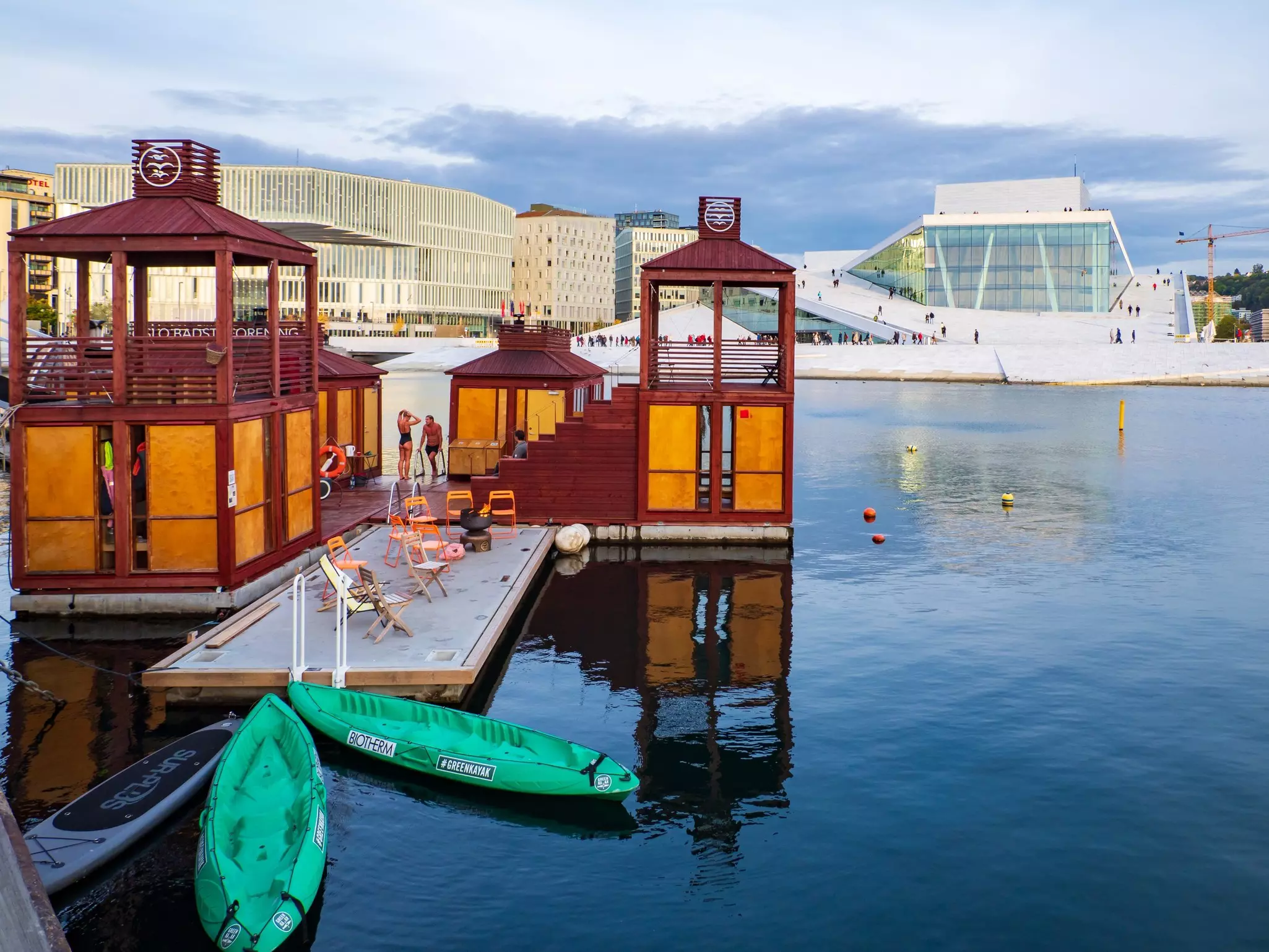 Green kayaks are pulled up to a dock with folding chairs that's part of a sauna complex in the harbor near the Oslo Opera House.