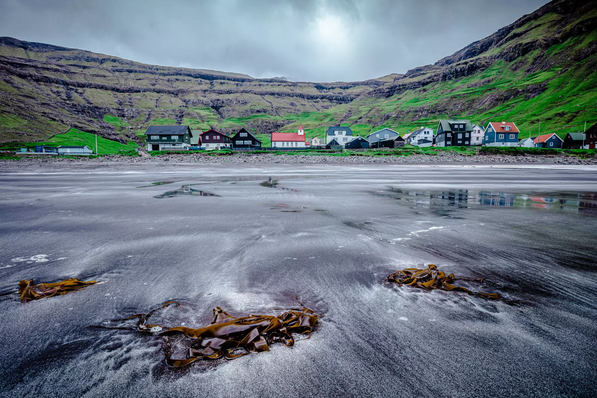 A black-sand beach backed by a small settlement with a red-roofed church, surrounded by hills.