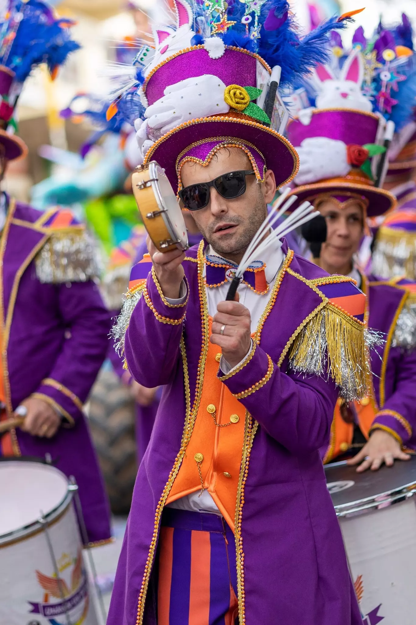 Man in parade wearing dark sunglasses and a purple and orange fringed, long jacket and purple whimsically decorated top hat shaking a tambourine and looking toward the camera