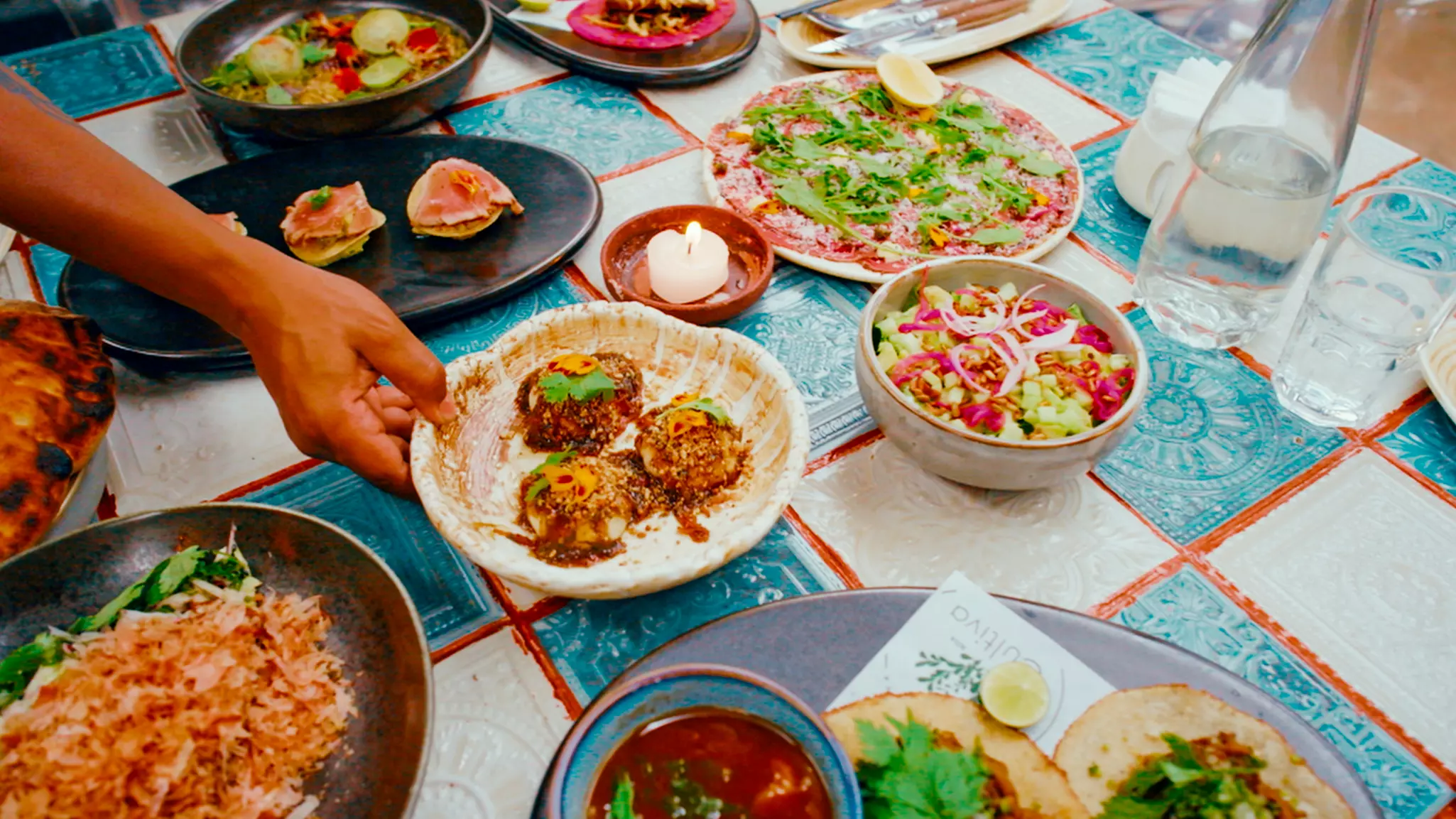 A series of dishes is seen from above, laid out on a plaid tablecloth. A person’s hand grabs one of the dishes.