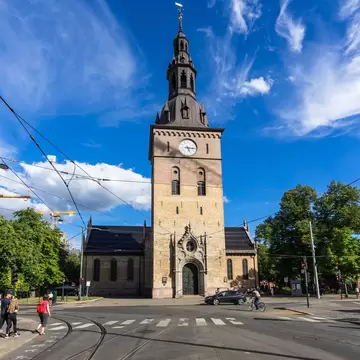 Brick church tower with a clock and dark spire above a tram junction in central Oslo, pedestrians crossing below