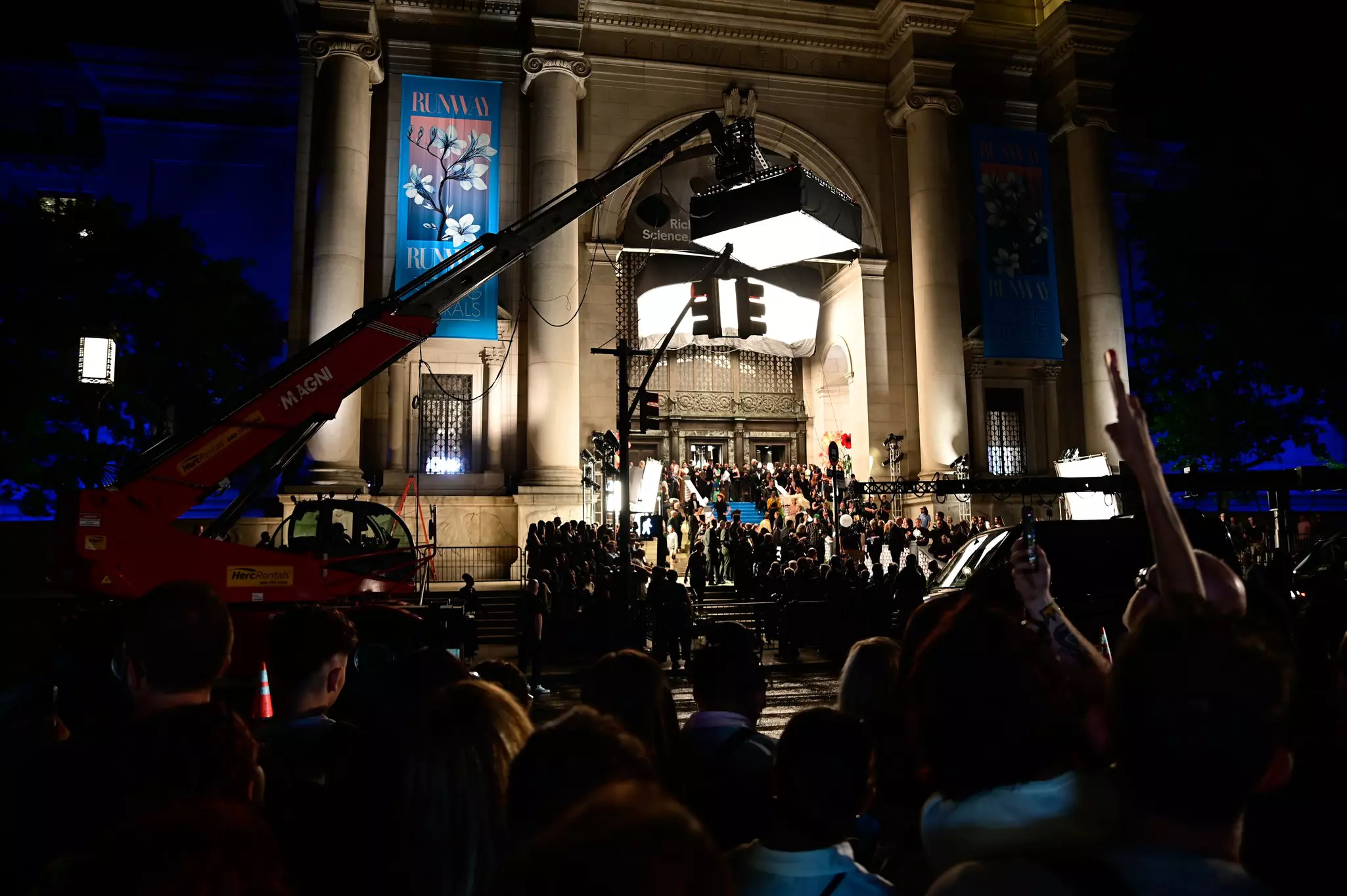 Film equipment is set up in a city street for a nighttime shoot in front of the entrance to a museum.