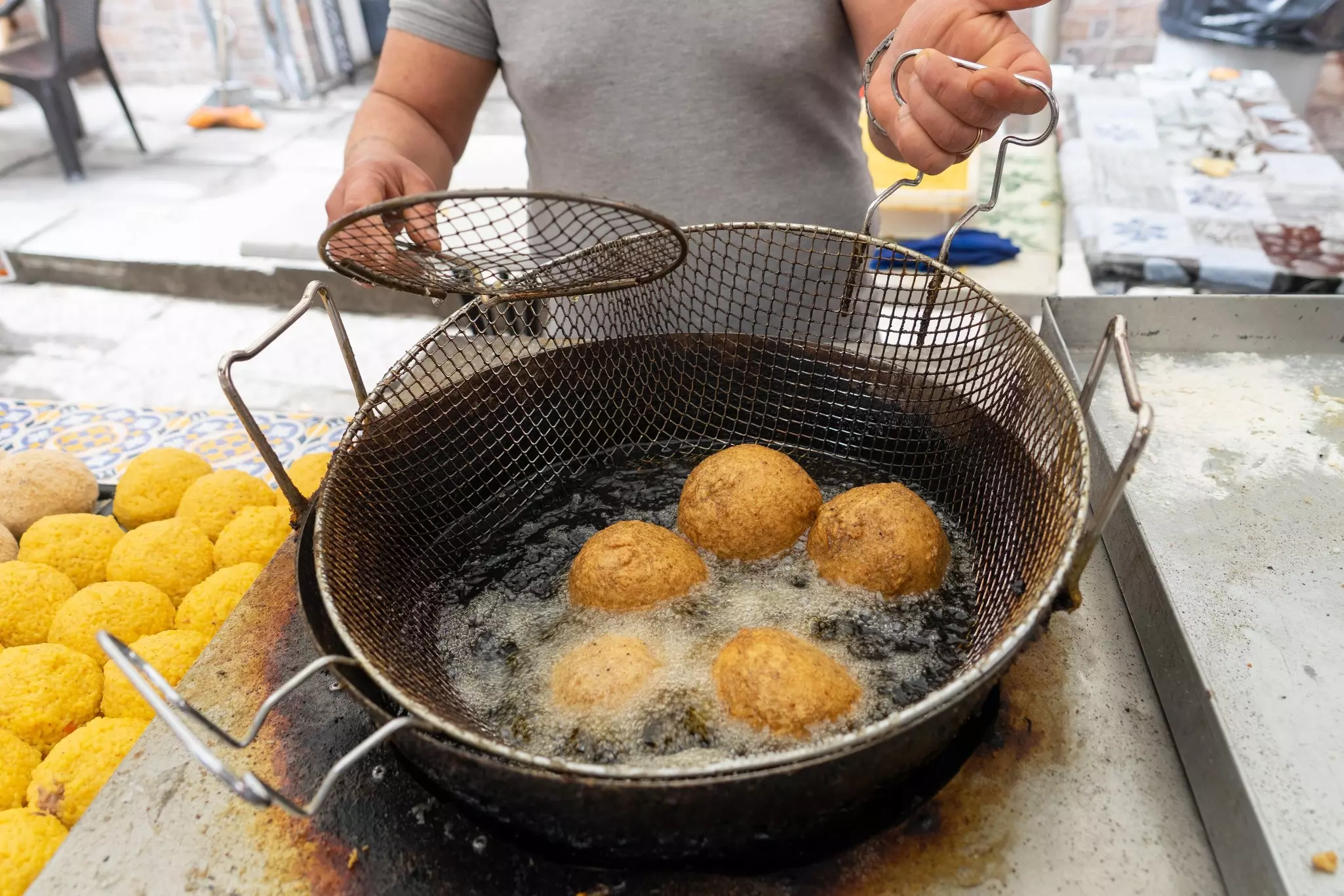 Five rice balls fry in oil; the vendor holds a strainer, and prepared balls waiting to be fried are to the left.