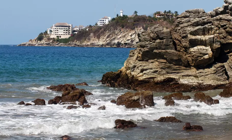 Surf washing over dark rocks below a sandstone outcrop in Puerto Escondido, hotels and a white lighthouse on the headland