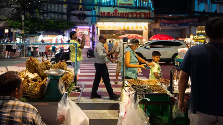 A mother and child and other pedestrians walk along a street in Bangkok with fruitsellers in the foreground. 