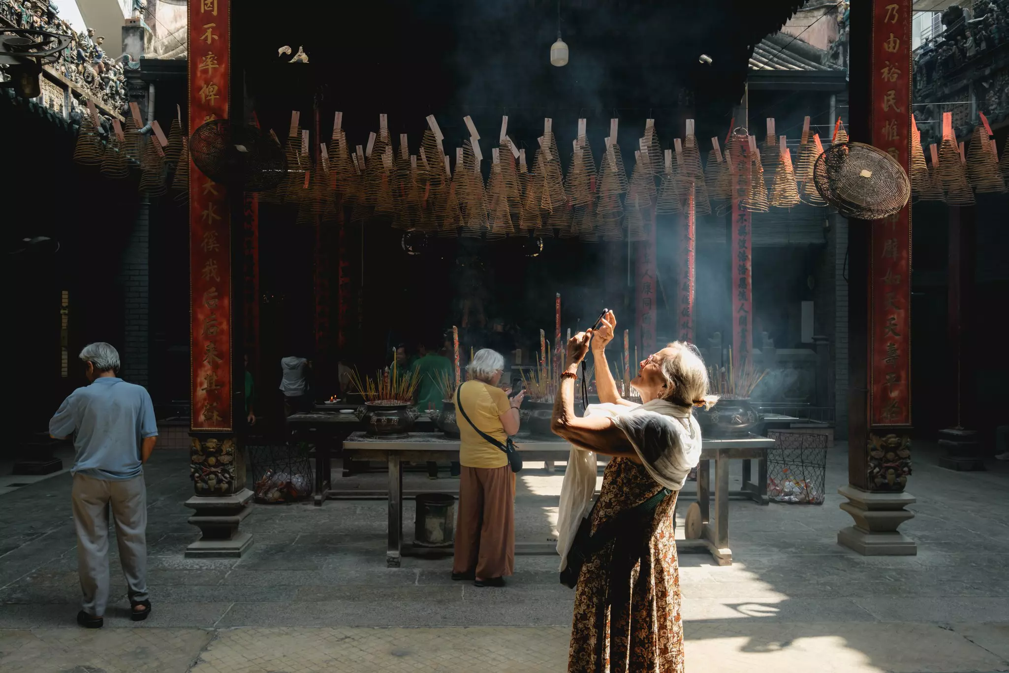 A woman takes a photo with her phone in a temple. Ornaments hang from the ceiling next to columns, and the smoke from incense is pictured in rays of sunlight.