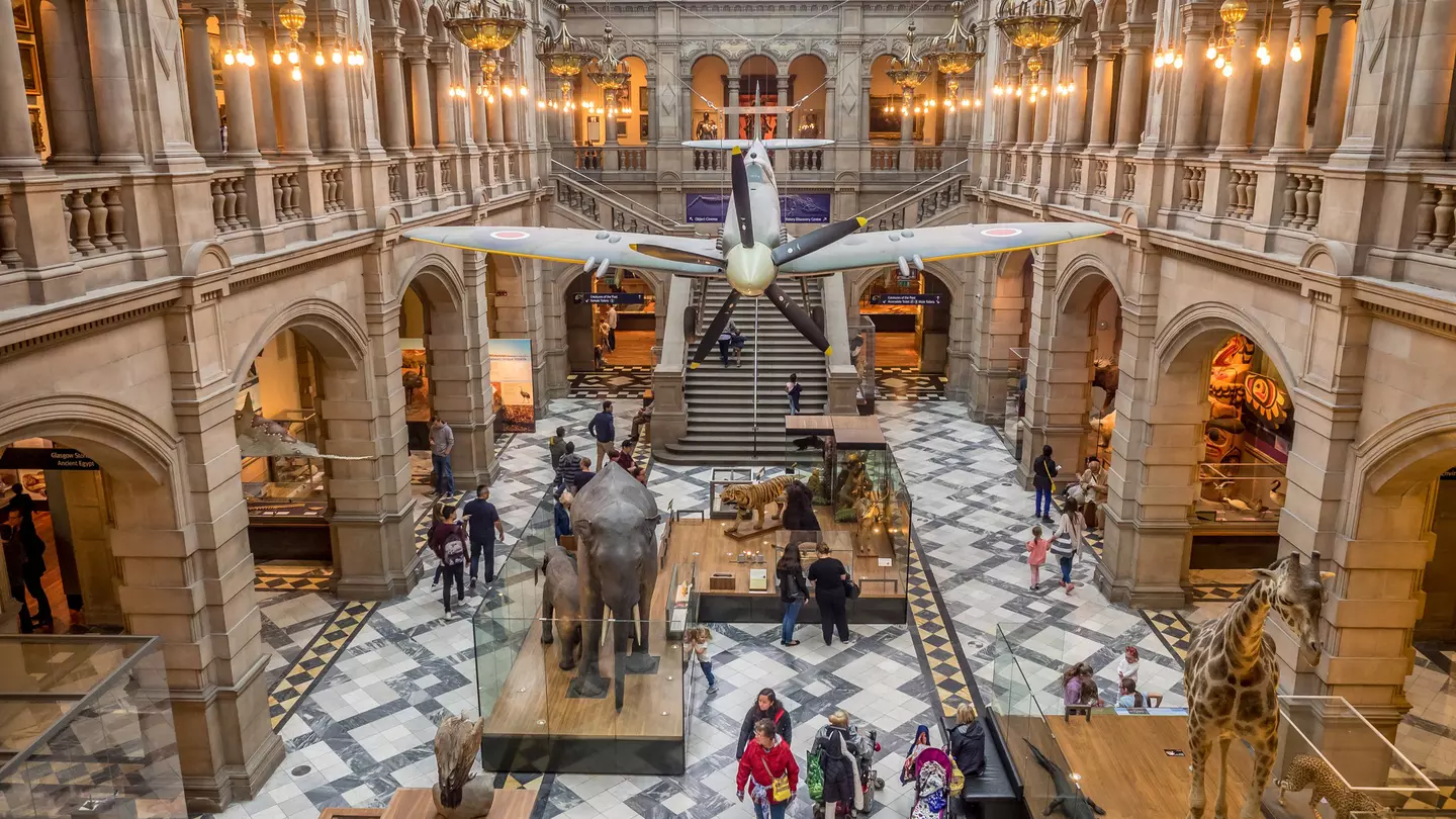 People walk below an airplane hanging in a large atrium with a black-and white floor in a museum in Glasgow, Scotland.