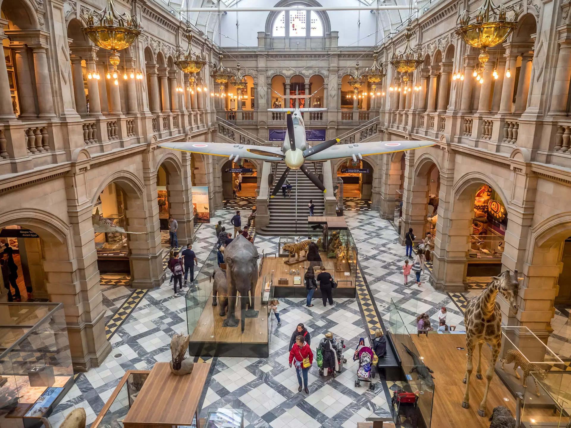 People walk below an airplane hanging in a large atrium with a black-and white floor in a museum in Glasgow, Scotland.